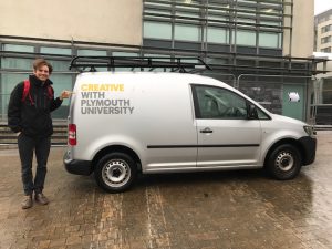 Oliver Broadbent stands next to a van that has the caption on it Creativty with Plymouth Univesity. Oliver is pointing at the word creativity. He was invited to the university to talk about teaching conceptual design