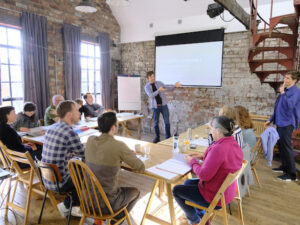 Image to advertise our How to have ideas course, showing Oliver Broadbent delivering a workshop to a group of 12 people sitting in a horseshoe shape in a sunlit training room
